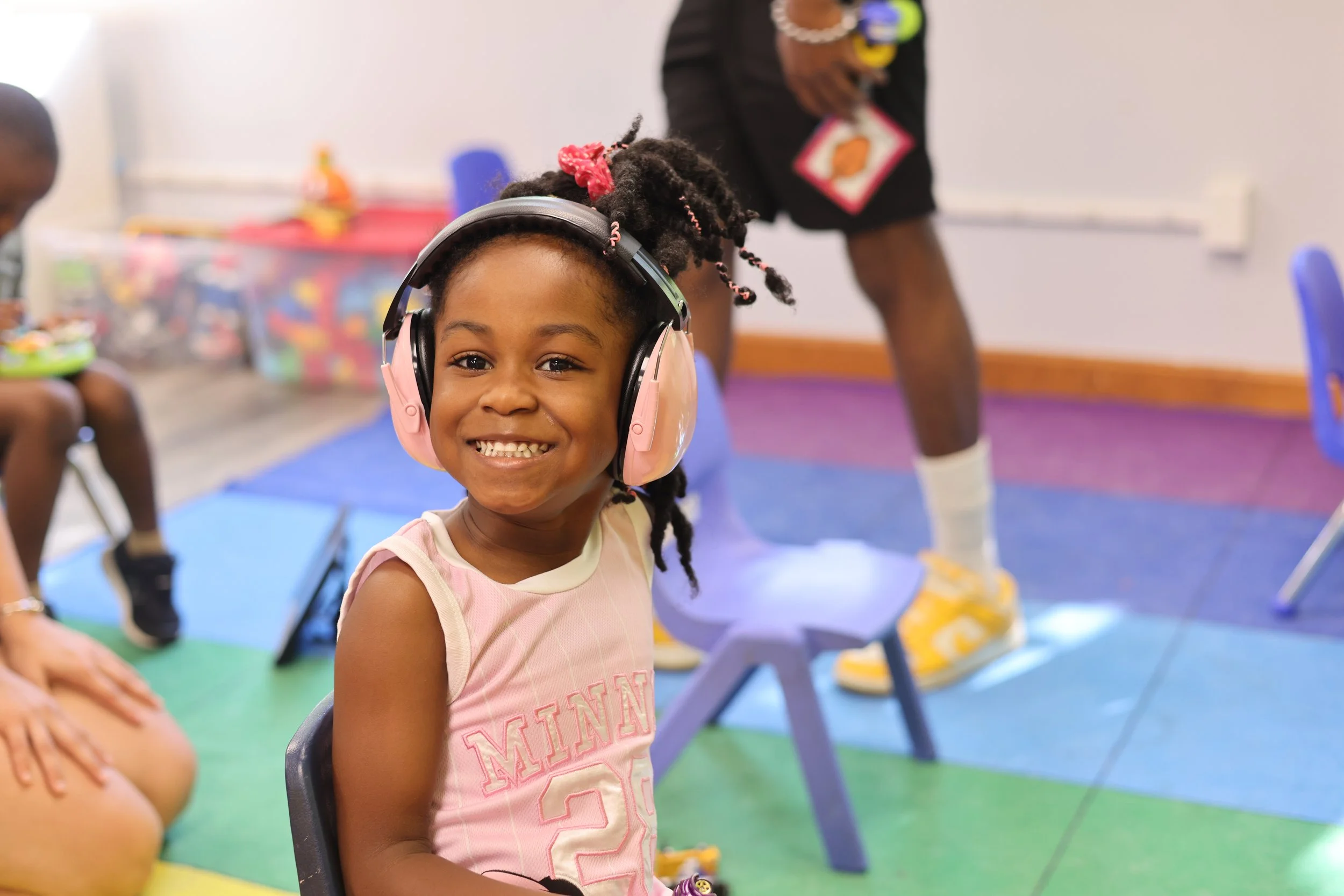 A young girl with braided hair, smiling, wearing pink headphones and a pink sleeveless shirt, sitting on a chair in a colorful classroom.