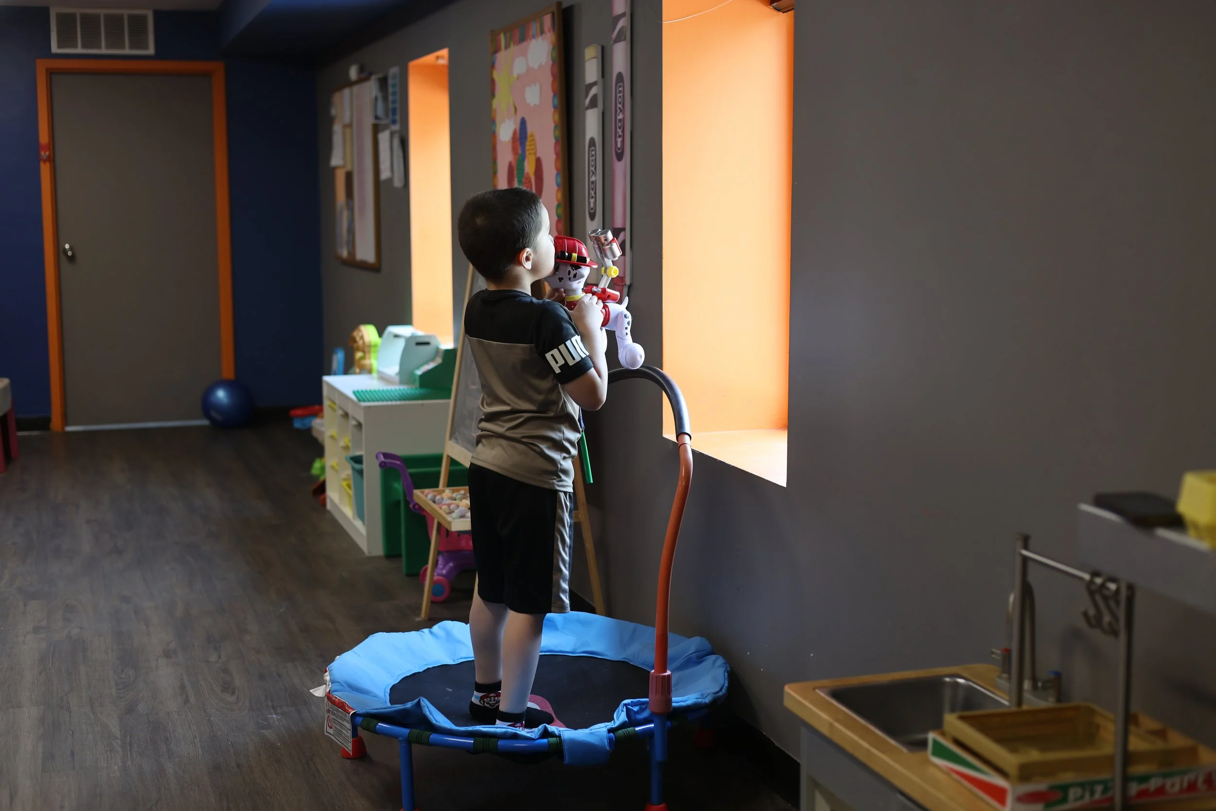 A boy standing on a small trampoline in a playroom, looking out a window and holding a fire truck toy.