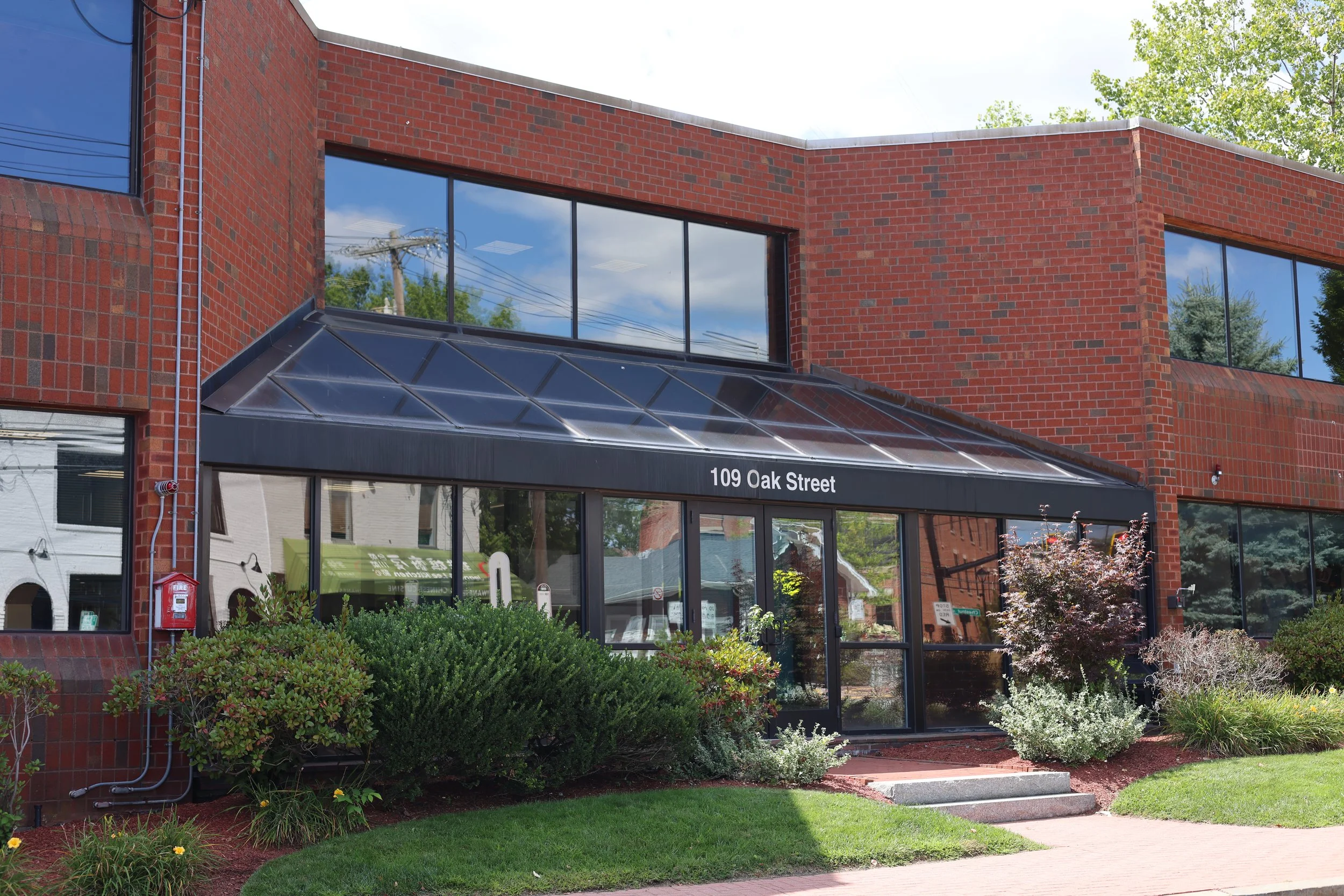 Brick commercial building with large glass windows, a black awning with the address 109 Oak Street, and landscaped plants and shrubs in front.