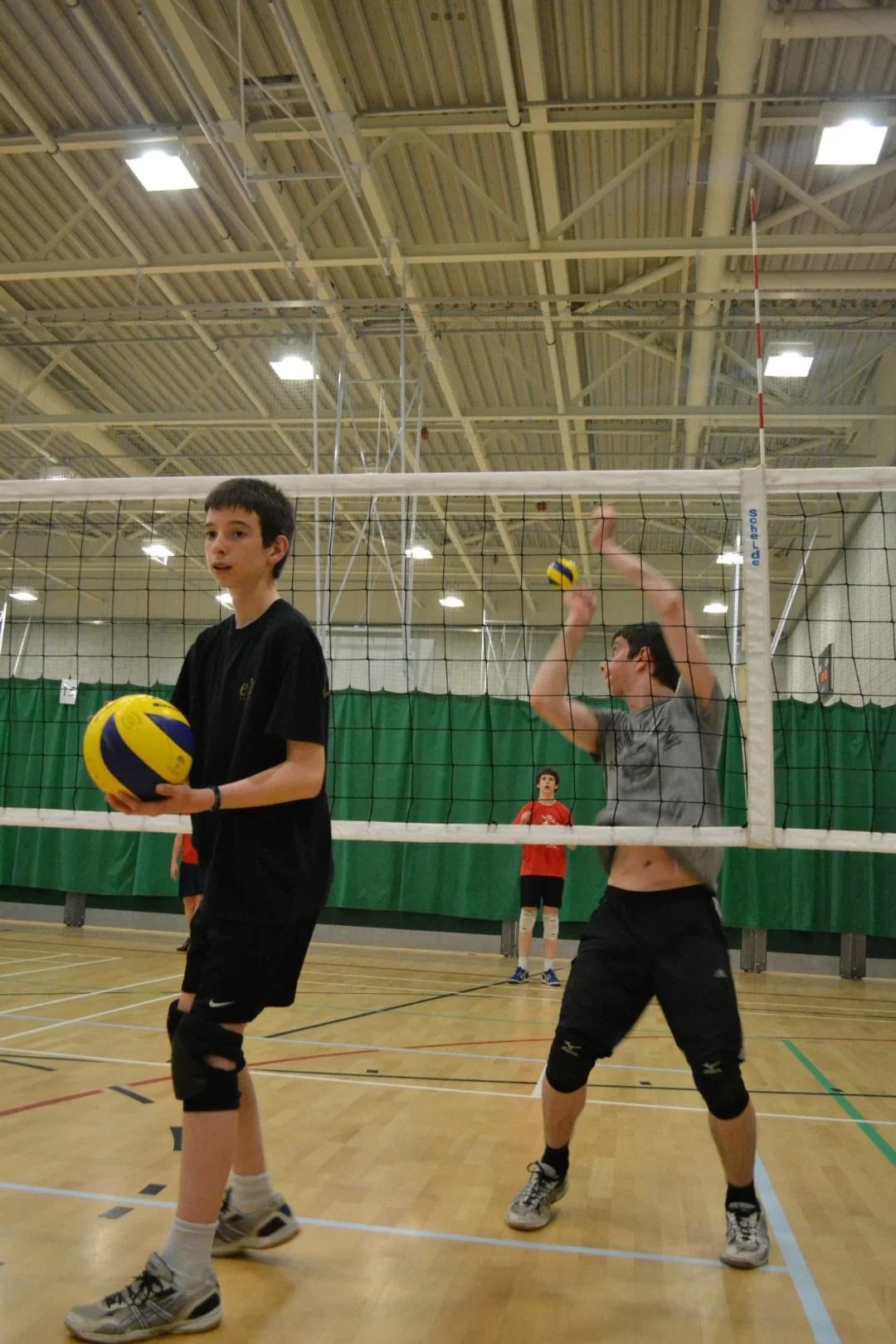 Two boys playing volleyball inside a gymnasium, one boy in a black shirt holding a volleyball, the other boy in a gray shirt preparing to hit the ball at the net, with a third boy in red in the background watching.