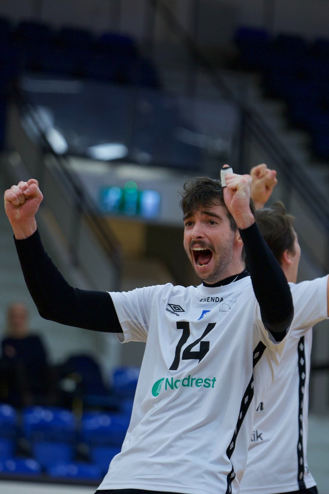 A male volleyball player wearing a white jersey with the number 14, celebrating with his arms raised and fists clenched, in an indoor sports arena.