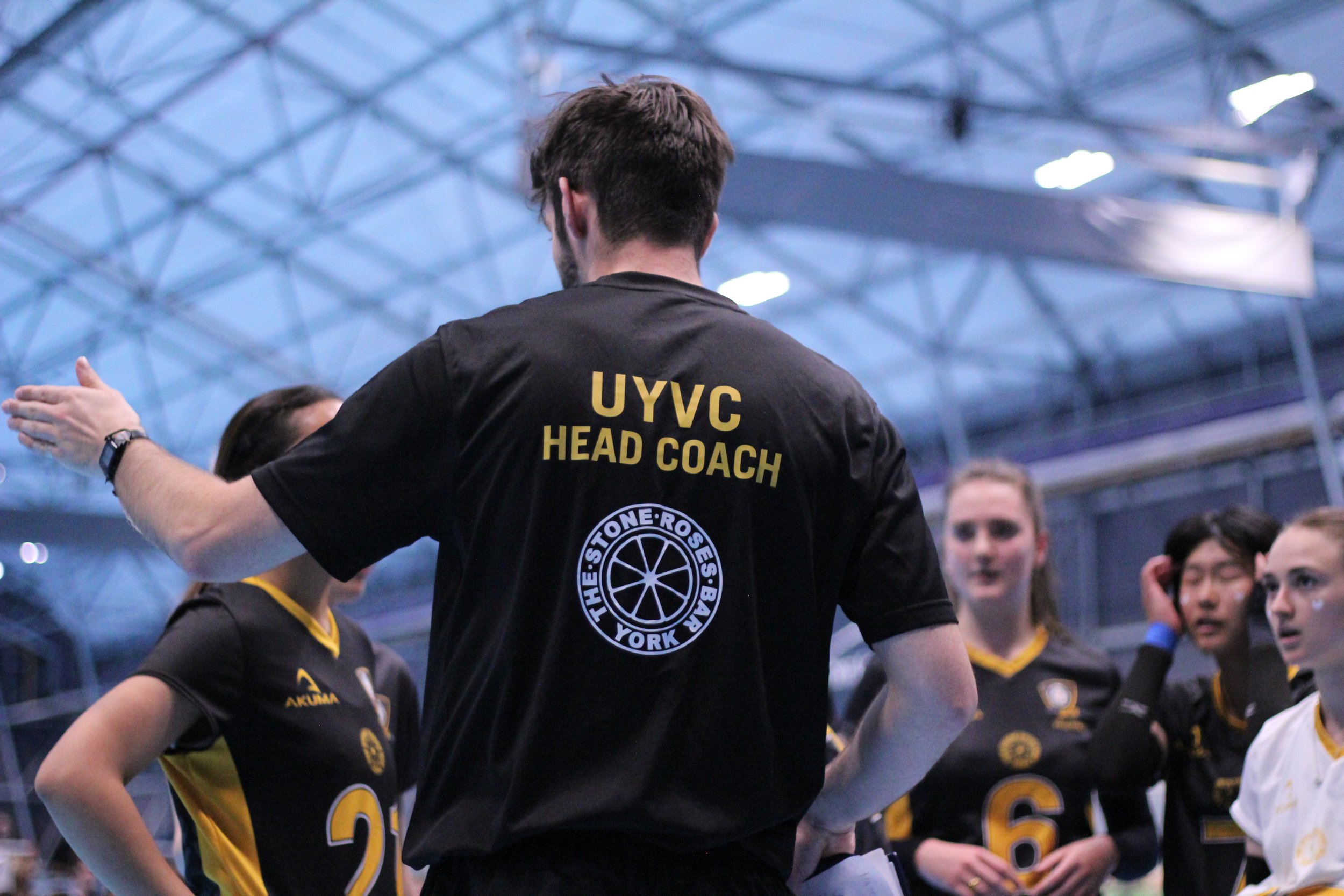 The head coach of the UYVC team giving instructions to female volleyball players in a sports facility.