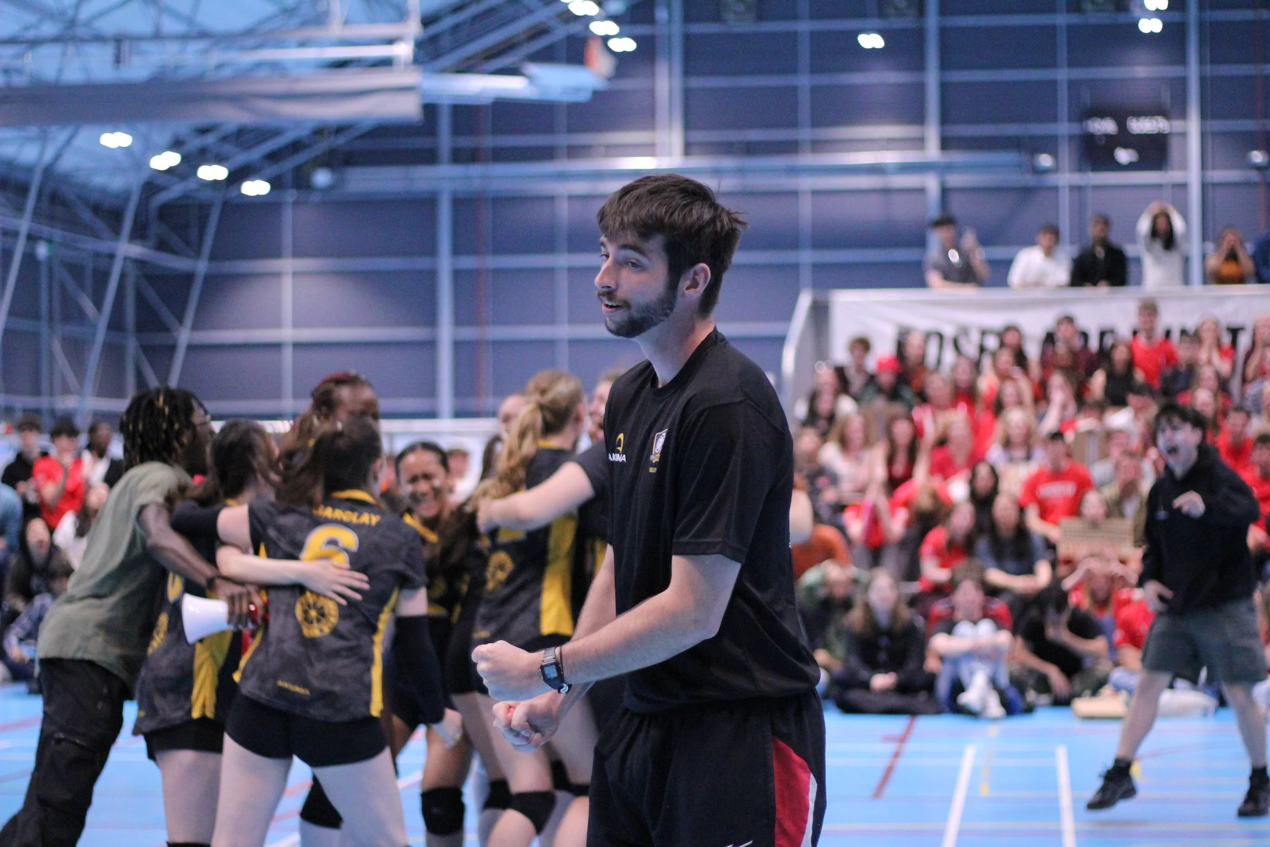 A group of young women in volleyball uniforms celebrating on an indoor volleyball court, with a man in a black shirt smiling in the foreground and spectators watching in the background.