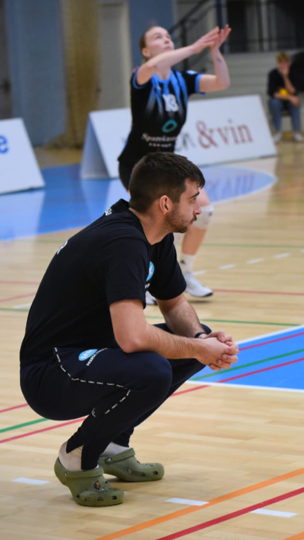 A man squatting on the sidelines of a sports court, watching a volleyball game, with a player preparing to serve in the background.