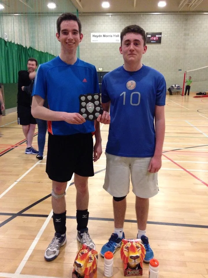 Two young men in blue sports shirts and shorts standing on an indoor gym court. One is holding a small trophy or award. There are other people in the background and a sign that says 'Haydn Morris Hall'.