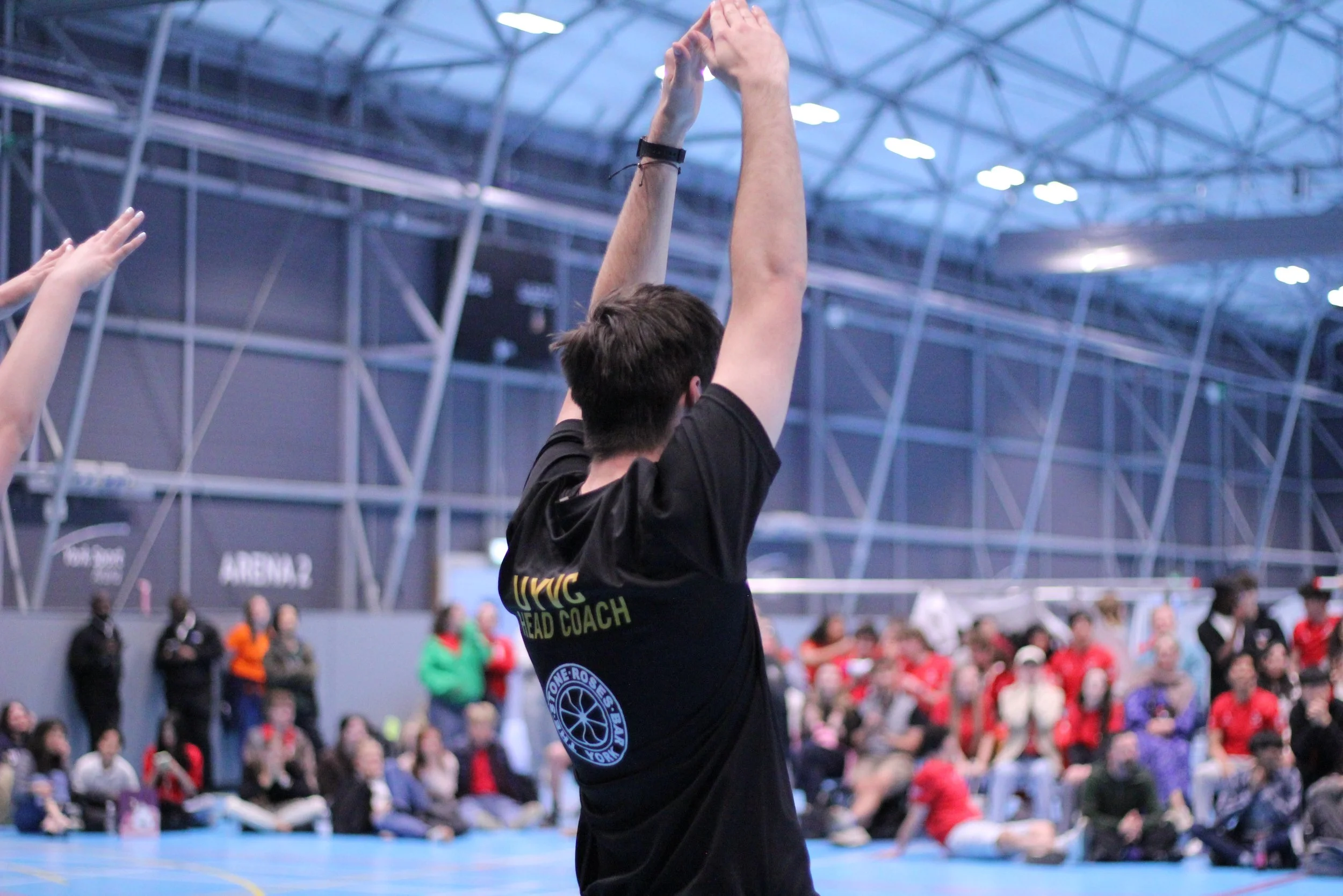 A person with short dark hair, wearing a black t-shirt labeled 'UWC HEAD COACH,' standing with arms raised in a large indoor sports arena filled with spectators, some wearing masks, while others watch and cheer.