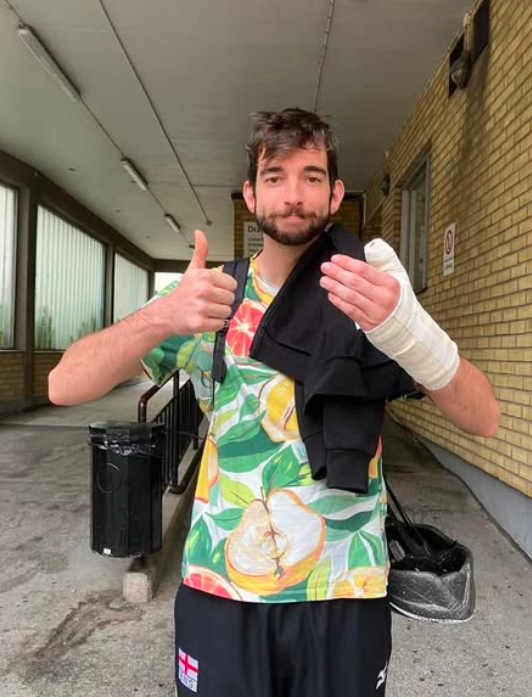 A man with a bandaged left arm giving a thumbs-up while holding a black jacket over his shoulder, standing outdoors near a brick building, wearing a colorful floral shirt and black shorts with a small England flag patch.