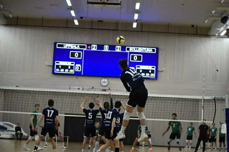 Indoor volleyball game with players at the net, some jumping to block the ball, opponents on the other side, and a digital scoreboard above.