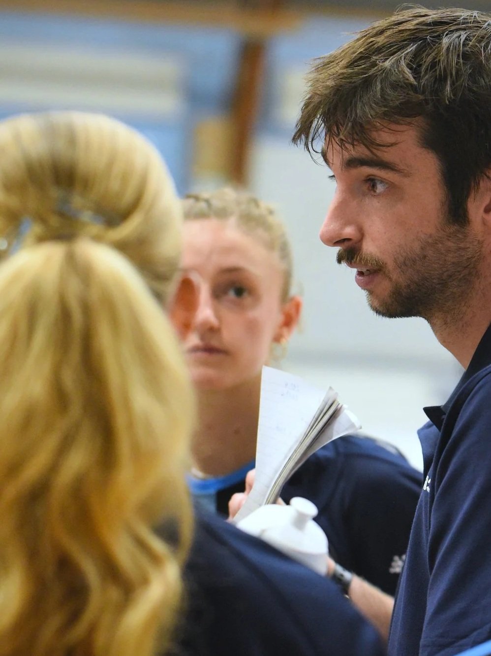 Three people having a conversation indoors, with a woman with long curly blond hair in the foreground and a man with dark hair and beard in profile on the right. A young woman with braided blond hair looks on in the background.