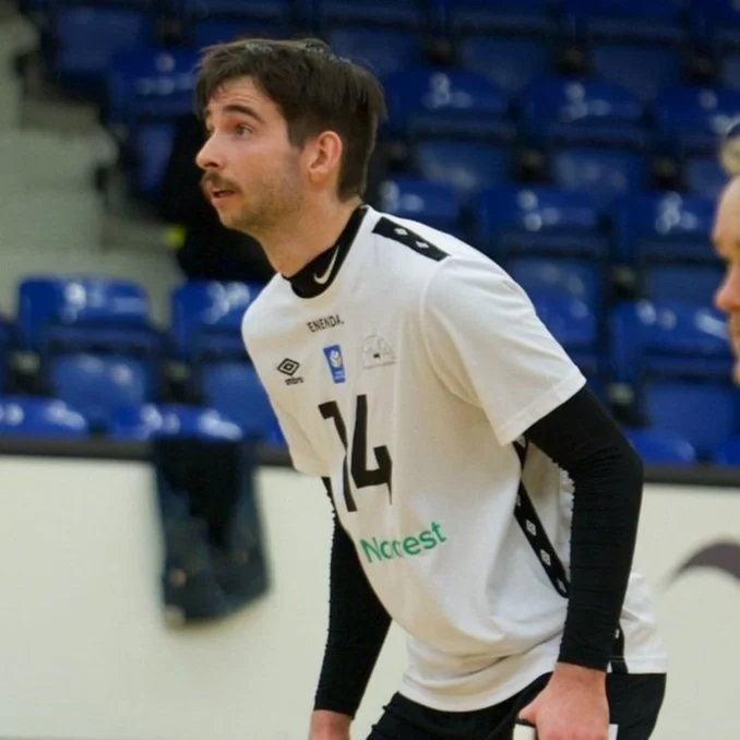 A male volleyball player in a white jersey with black accents, number 14, standing on an indoor court with blue seats in the background.