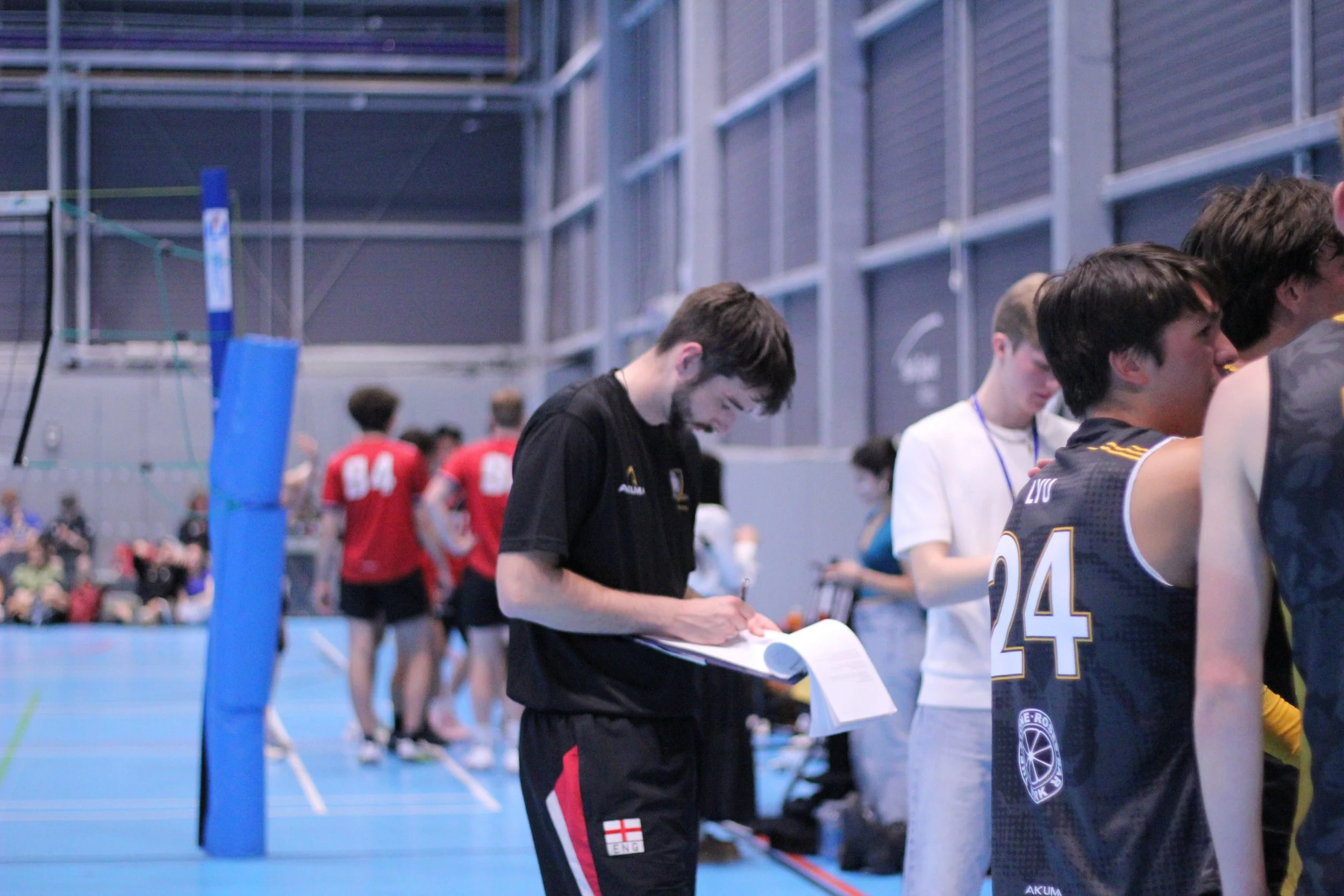 Young male volleyball players and officials on an indoor court, some wearing red uniforms with numbers, others in black, with a volleyball net in the foreground and spectators in the background.