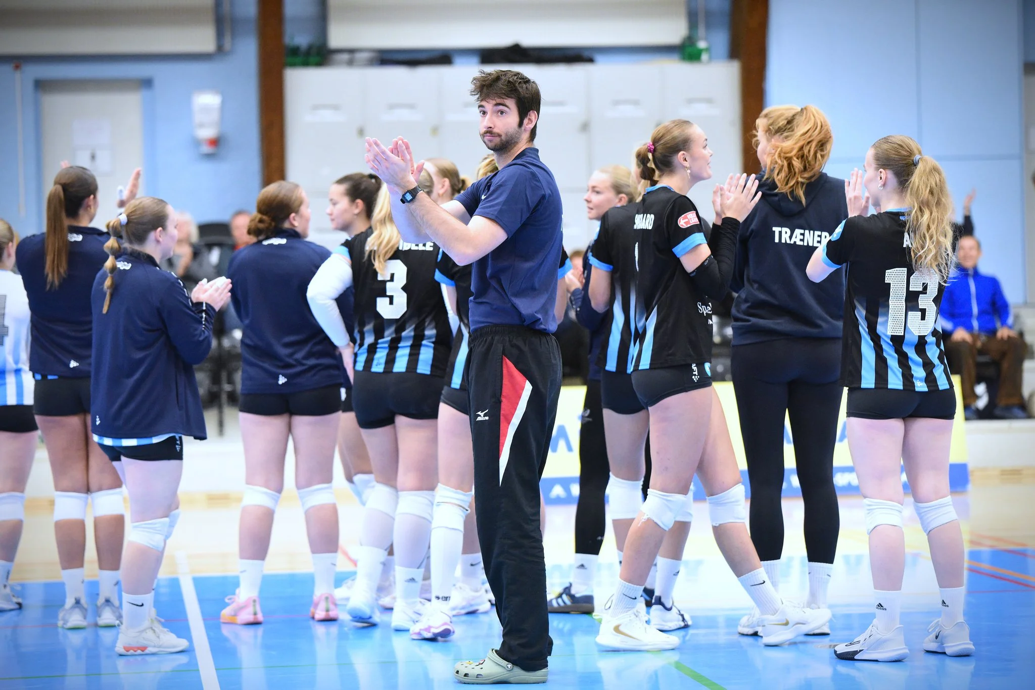 A women's volleyball team in black and blue jerseys clapping and celebrating during a match, with a male coach standing in the center.