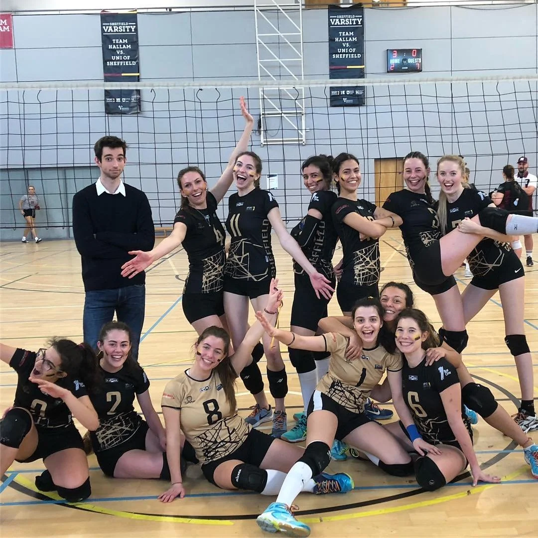 A girls' volleyball team celebrating on the court after a match, with some players sitting on the floor and others standing behind them, all smiling and making joyful gestures.