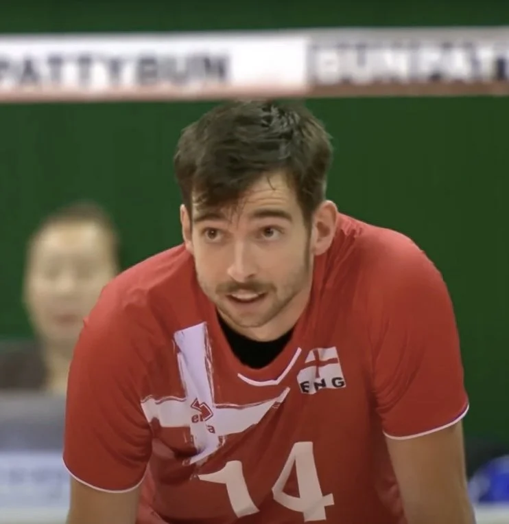 A man in a red sports jersey with the number 14 and England's flag emblem, leaning forward on a sports court during a volleyball match.