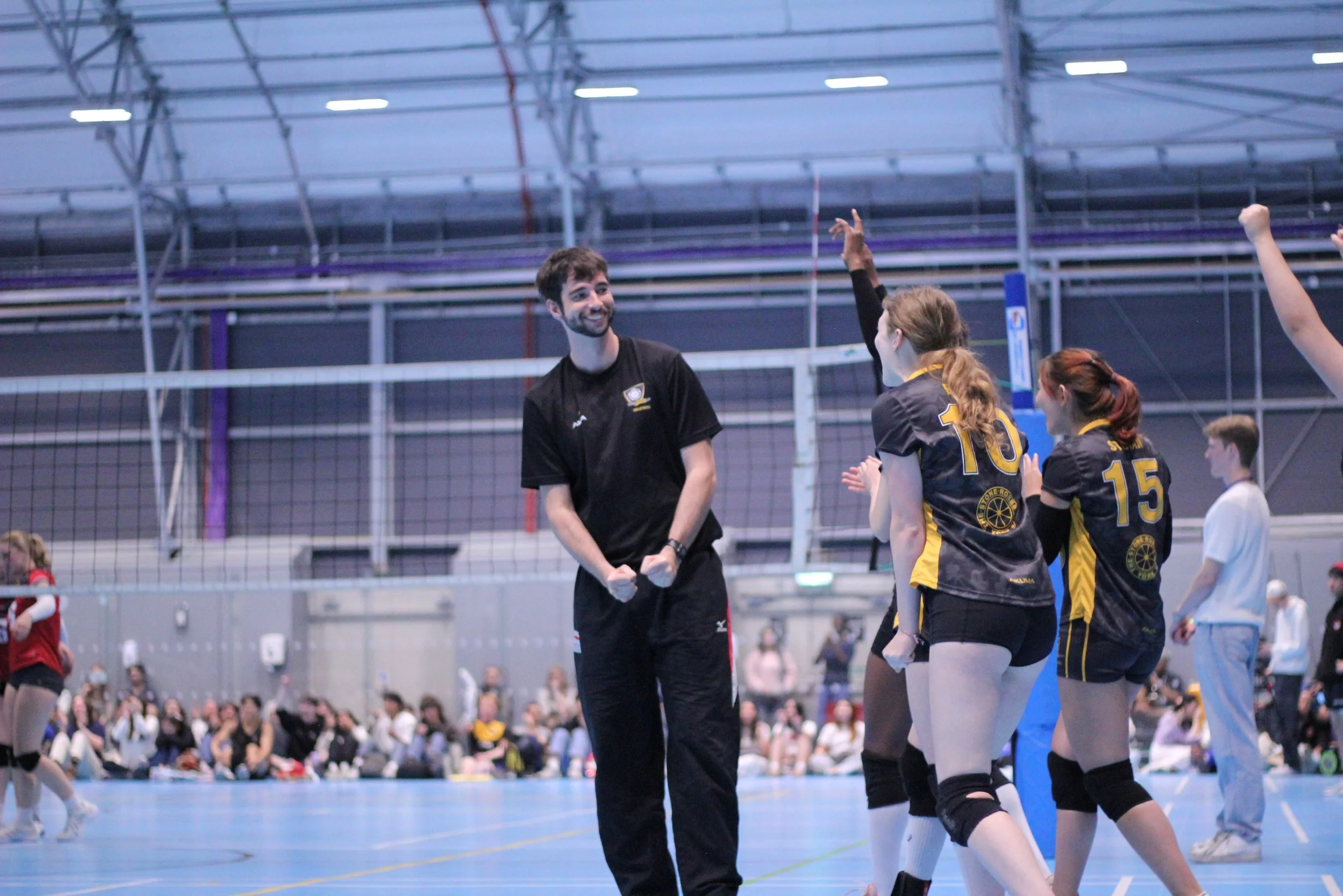 A volleyball coach or  smiling at a team of female volleyball players in black and yellow uniforms during a match in an indoor gymnasium. The players are celebrating or cheering, and one player has her fist raised. Spectators are visible in the background.