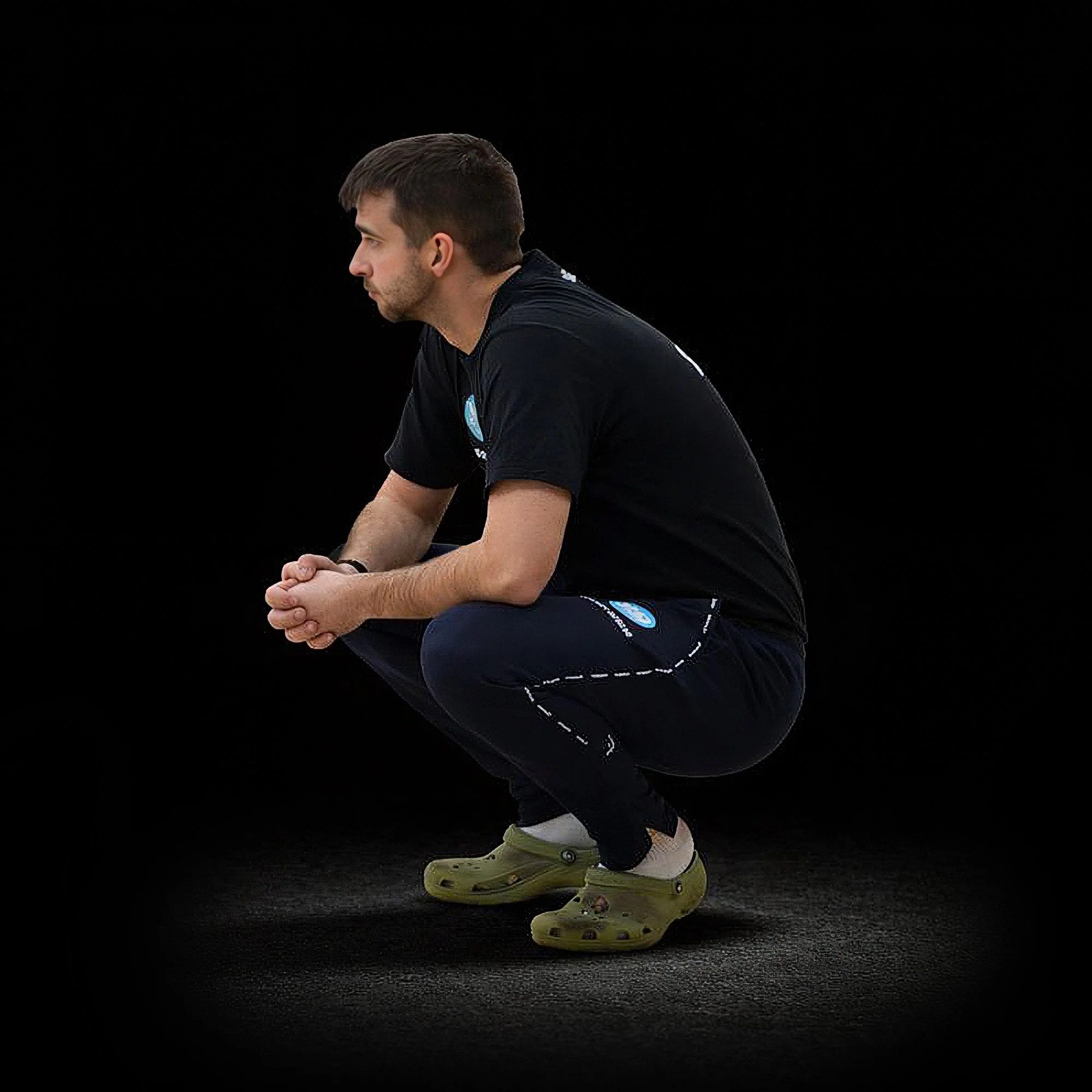 A man in black clothing and green Crocs squatting with hands clasped against a dark background.