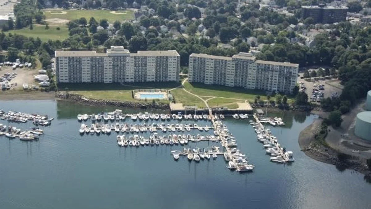 Aerial view of a marina with numerous docked boats, a large apartment complex, a swimming pool, parking lot, and surrounding green landscape.