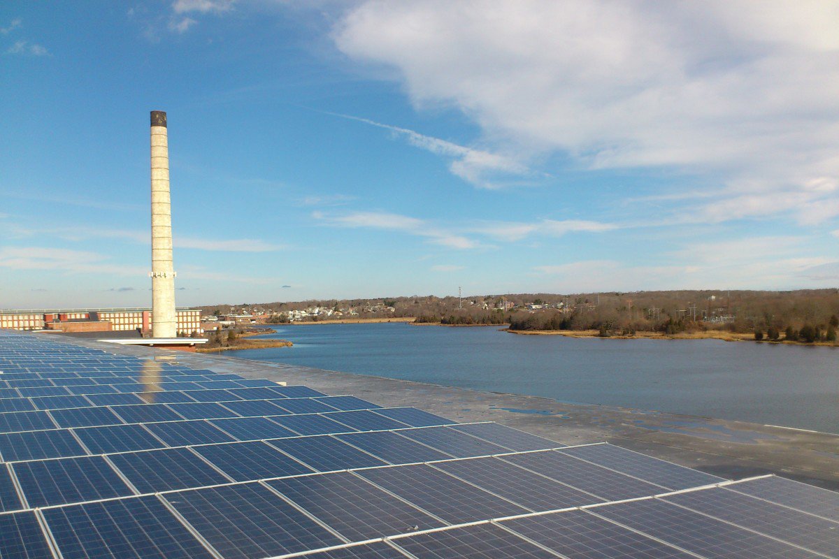 Solar panels installed on rooftop near a body of water, with a tall industrial chimney, in a partly cloudy sky.