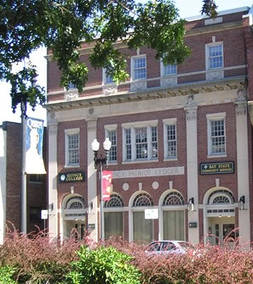 Historic brick building with banners for college and bay state, featuring three arched entrance doors, decorative columns, and windows, surrounded by greenery and trees.