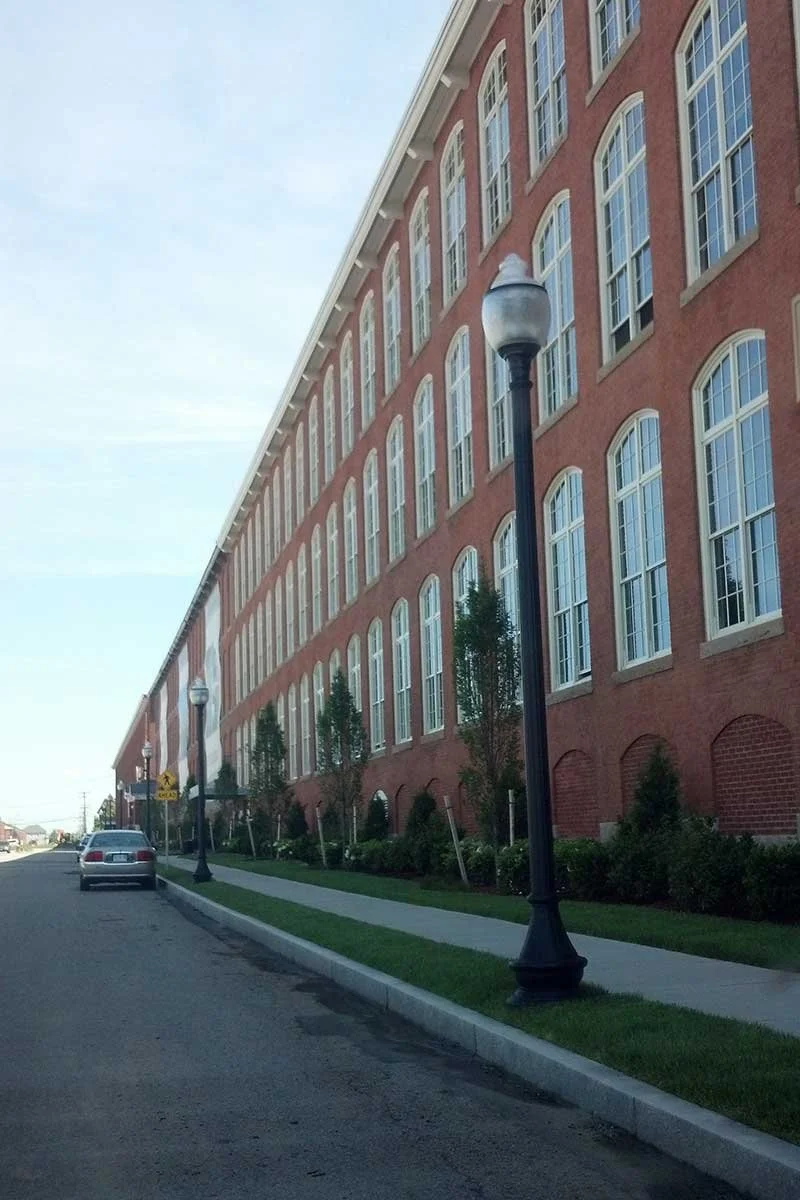 A large red brick building with multiple windows, sidewalk, street lamps, and parked cars on the side of the road.