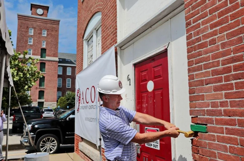 A construction worker wearing a white hard hat is chipping away at a red door with a tool in front of a brick building, with a sign that reads 'Keep Out' and a banner with the logo for ACO.