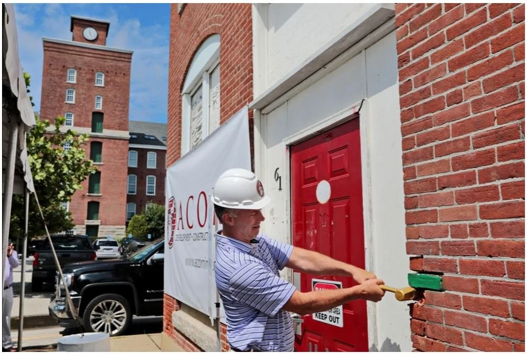 A man wearing a white construction helmet uses a hammer to break a brick wall in front of a red door. There is a banner with the logo of a construction company, and a parking lot with cars in the background.