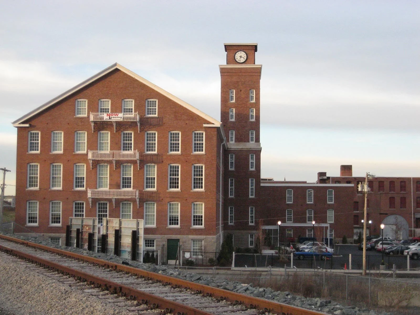 A brick building with a clock tower, parking lot, and train tracks in the foreground.