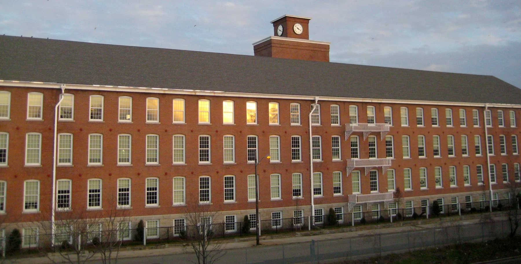A large brick building with multiple windows and a clock tower on top, illuminated by the setting sun.