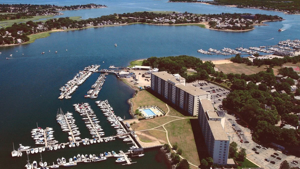 Aerial view of a marina with numerous boats docked, adjacent to a large beachfront apartment complex, with a town and body of water in the background.