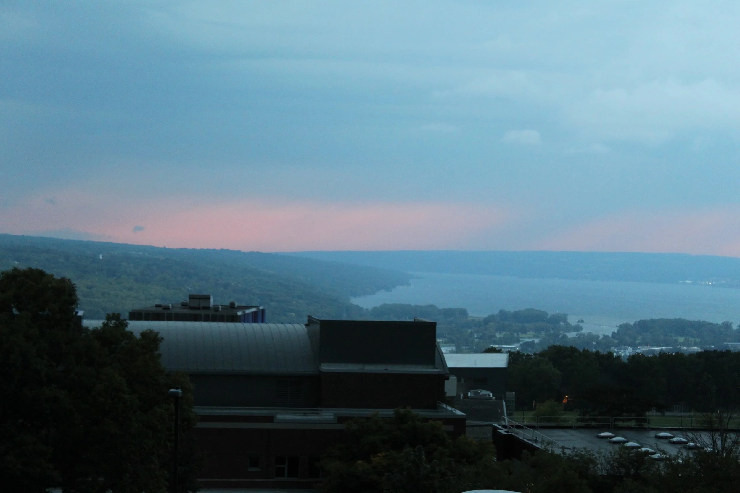 A scenic view of a river or lake with surrounding trees, a building with a curved roof in the foreground, and a pink and blue sky during dusk or dawn.