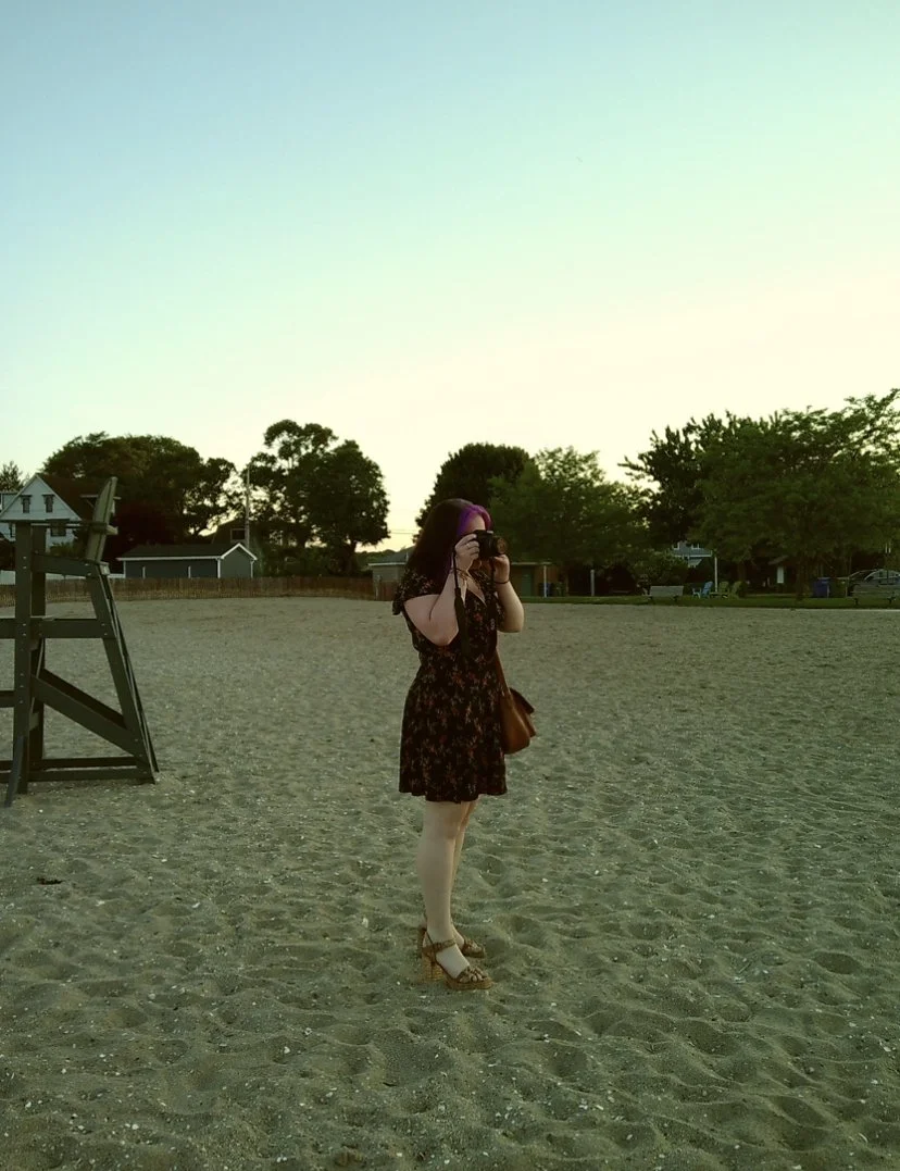 A woman wearing a black floral dress and high heels is taking a photo with a camera on a sandy beach during sunset.