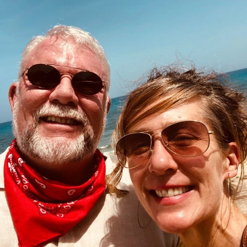 A smiling older man and woman taking a selfie on the beach with the ocean and blue sky in the background. Both are wearing sunglasses and the man has a red bandana around his neck.