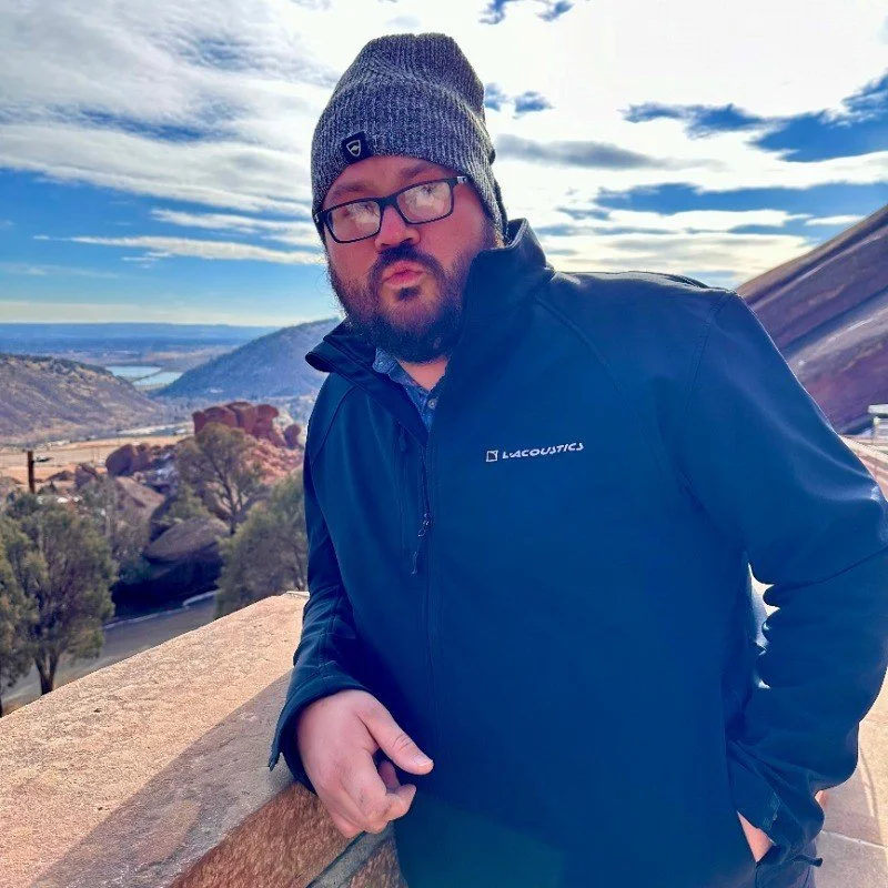 A man wearing a gray knit beanie, glasses, and a navy Lacoustics jacket is leaning on a stone railing outdoors. The background features a scenic landscape with rolling hills, sparse trees, and a river, under a partly cloudy sky.