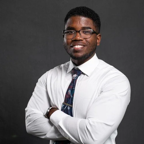 Professional portrait of a young man with glasses, wearing a white dress shirt, patterned tie, and a watch, smiling with arms crossed against a dark background.