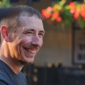 A man smiling outdoors with blurred colorful flowers in the background.