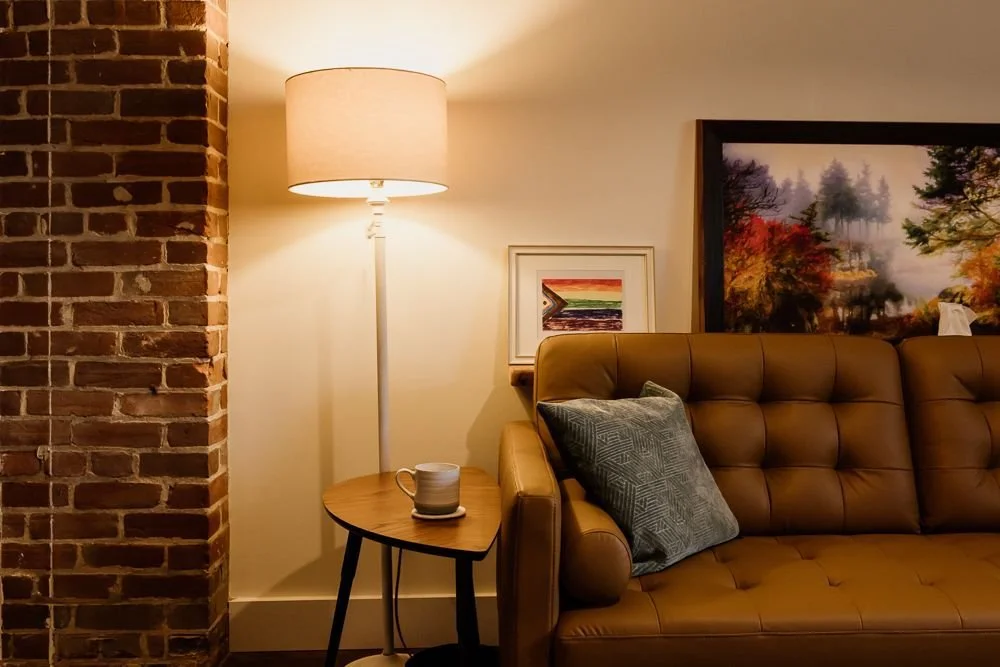 Living room with a brick wall, a floor lamp, a small wooden side table with a mug, a brown leather sofa with a throw pillow, and framed artwork on the wall.