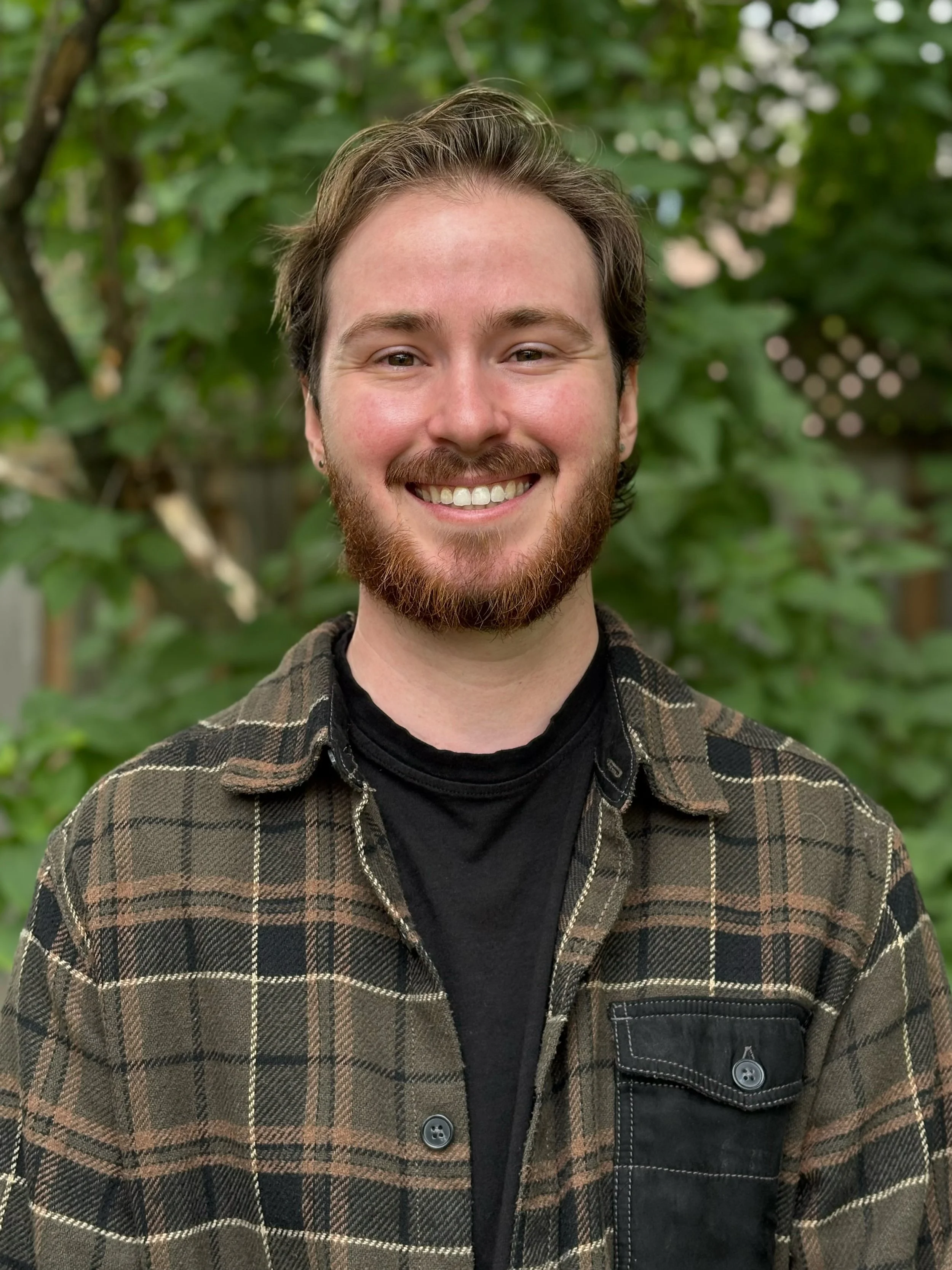 A smiling young masculine person with a beard and mustache, wearing a green plaid jacket over a black shirt, standing outdoors with green trees in the background.