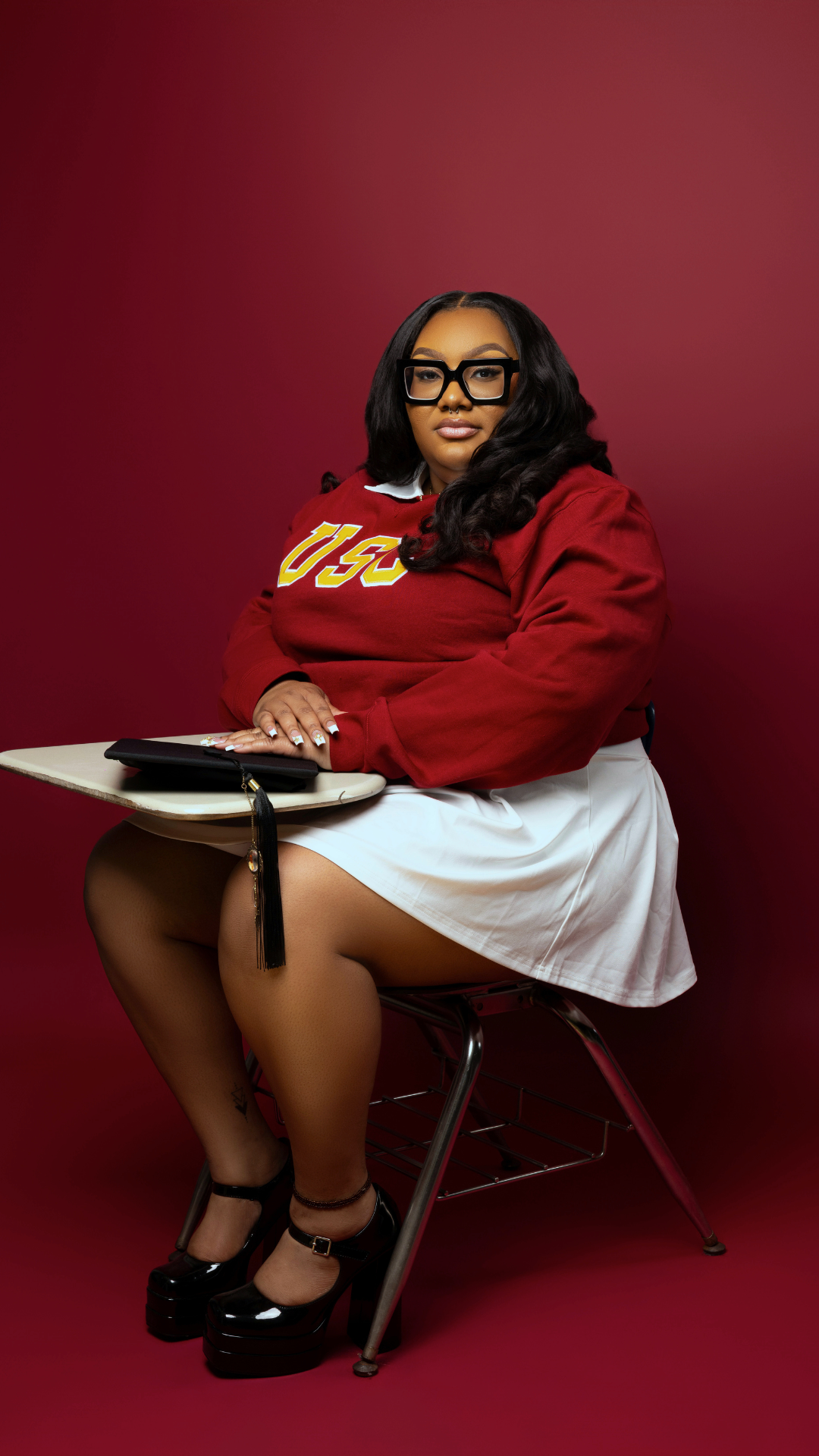 A woman with long black hair and large black glasses sitting on a chair with a red background. She is wearing a USC sweatshirt, white skirt, black platform heels, and has a small black handbag on her lap.