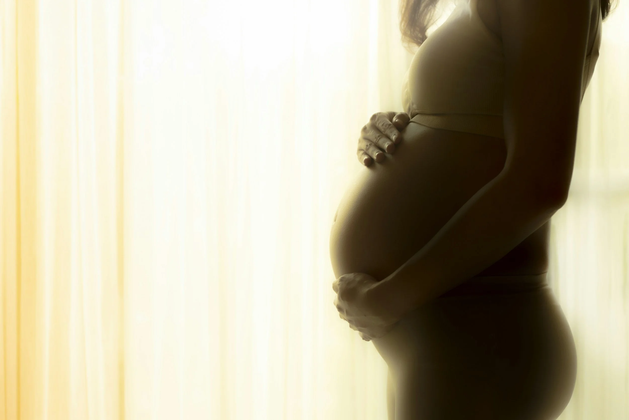 Close-up of a pregnant woman with her hands resting on her belly, standing in front of a softly lit curtain.