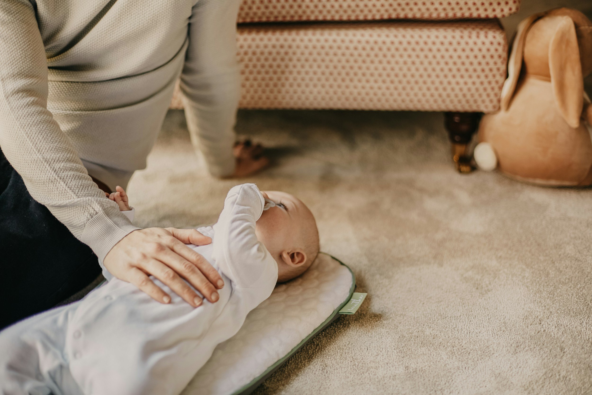 A baby lying on its back on a mat, being gently held and examined by an adult whose hands are visible. The adult is wearing a light-colored sweater, and the baby is in a white outfit. A couch with a red patterned cover and a ceramic dog figurine are in the background.