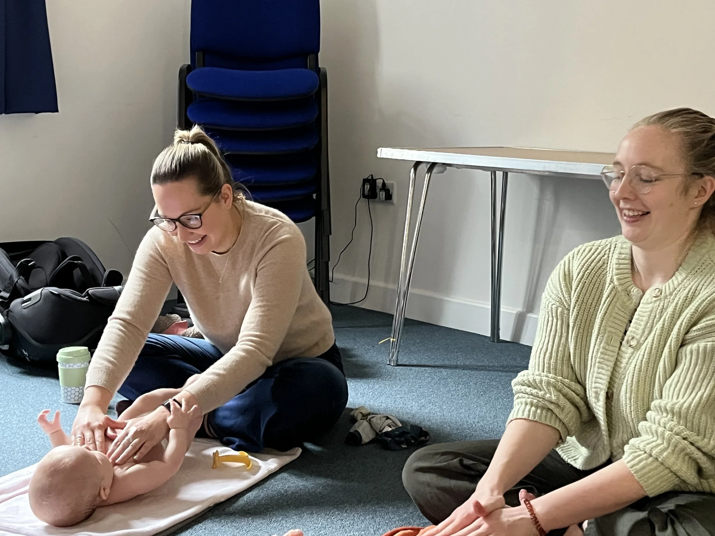 Two women sitting on the floor practicing baby massage in a baby massage class. One woman is wearing glasses and a beige sweater, the other has red hair and is wearing a green sweater. In the background, there are stacked blue chairs and a table.