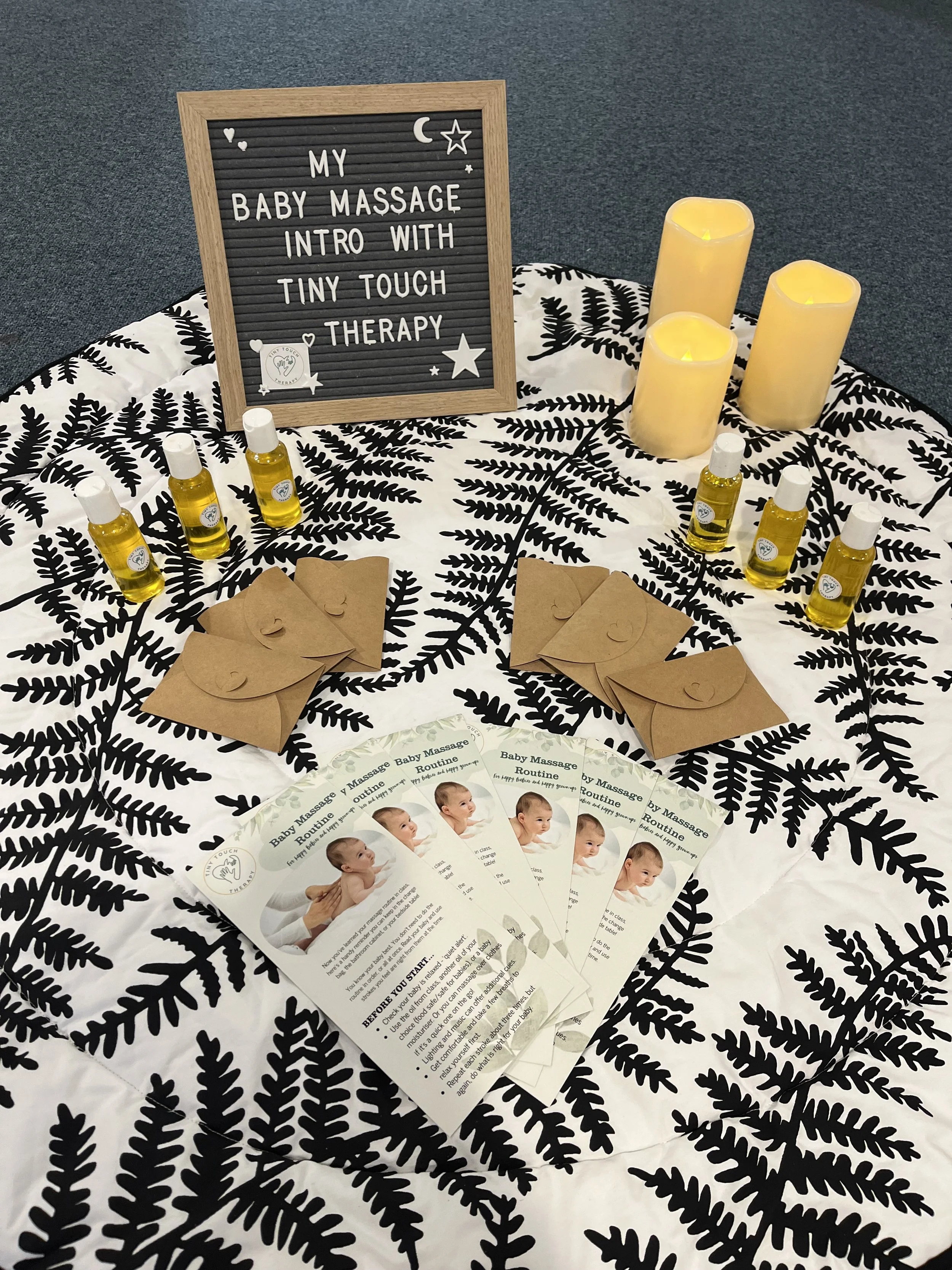 Display for a baby massage class with informational pamphlets, bottles of massage oil, decorative lights, a black felt letter board with white letters, and leaf-patterned tablecloth.
