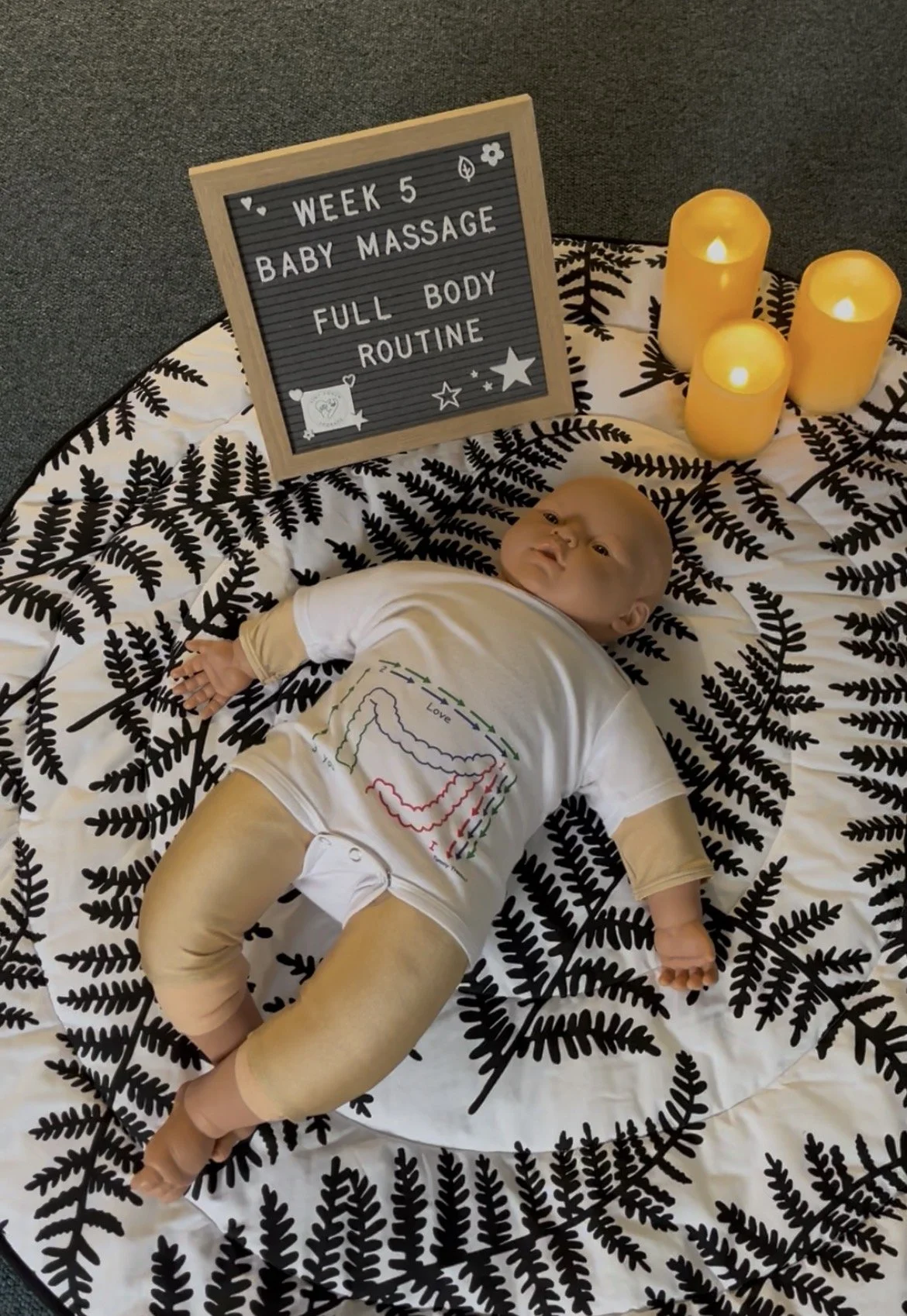 A baby lying on a black and white fern-patterned blanket surrounded by three yellow candles. There is a small board with the text 'Week 5 Baby Massage Full Body Routine' and some decorative stars and hearts.