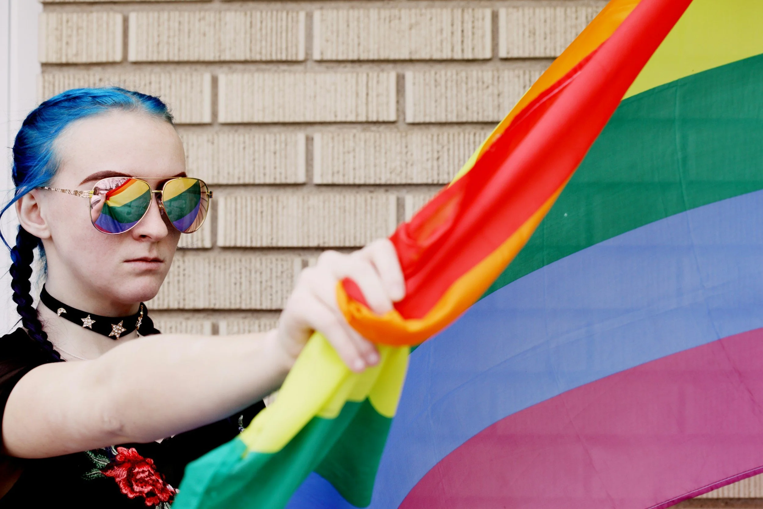 Woman with blue braided hair and stern expression is holding a rainbow flag in front of her as if to block and protect herself from danger