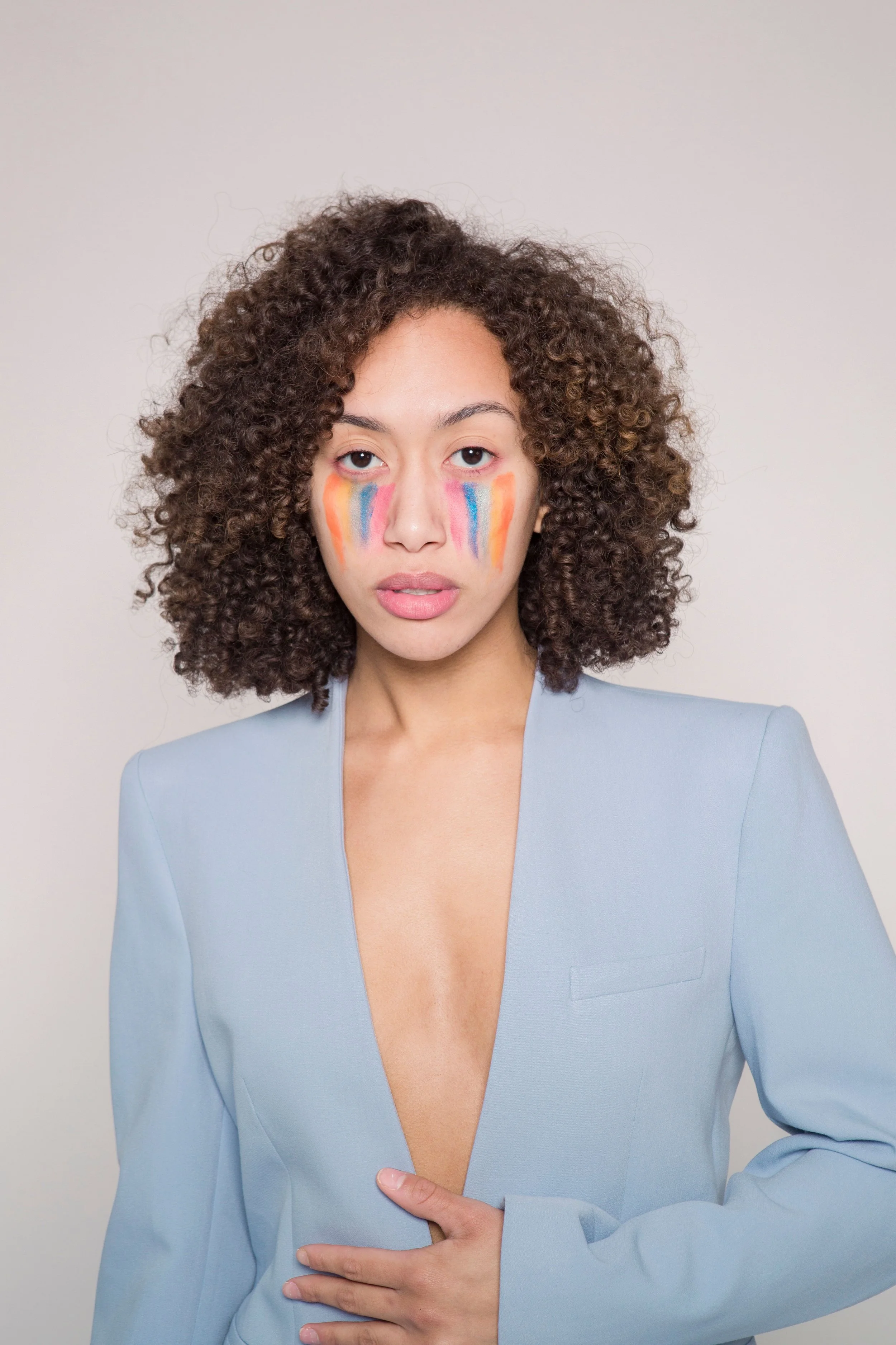 A person in a powder blue suit jacket without a shirt underneath, with brown coily hair and rainbow marks on the face, resembling tear marks.