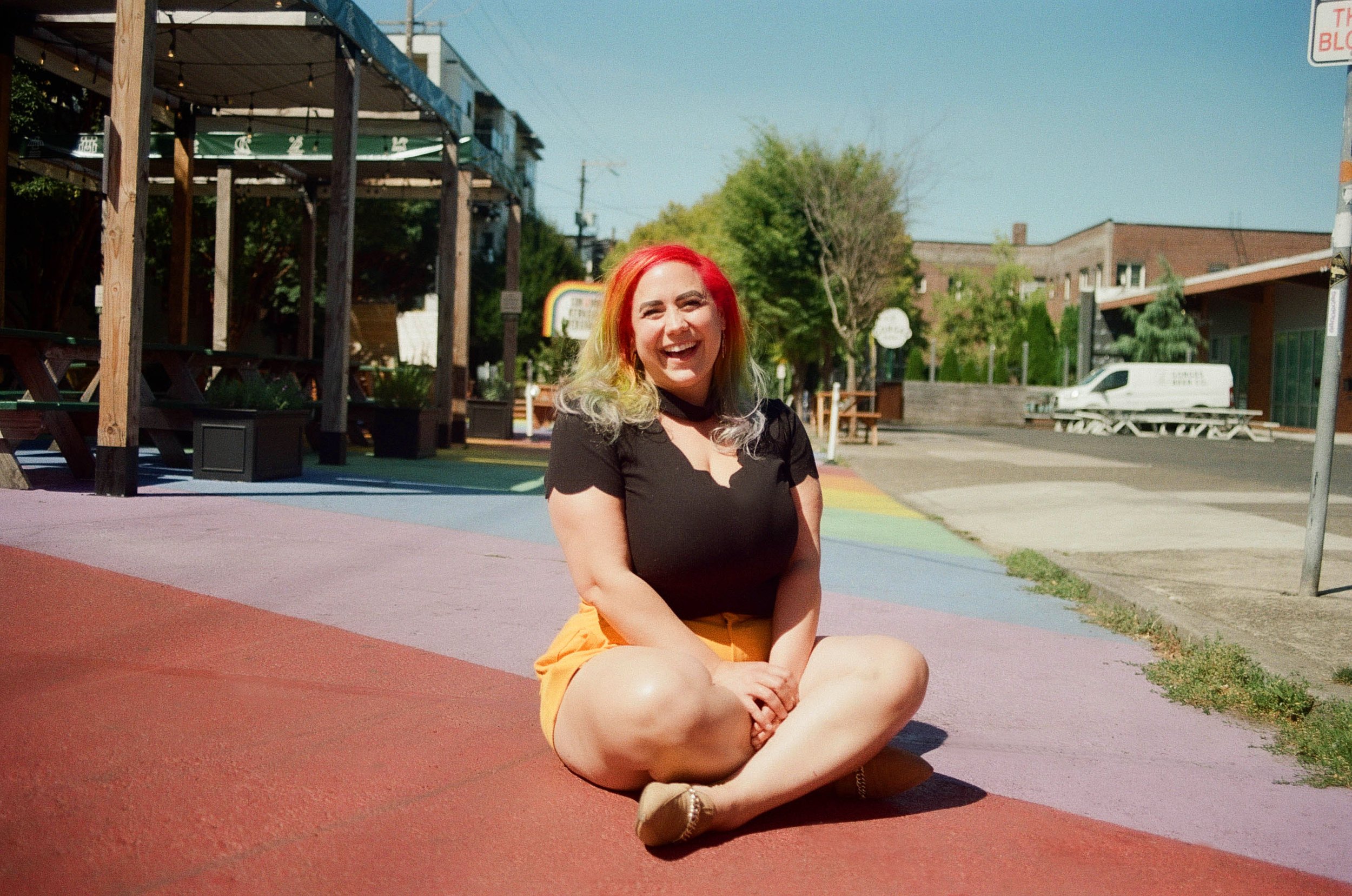 Woman with rainbow hair sitting cross legged and smiling, seated on a rainbow street