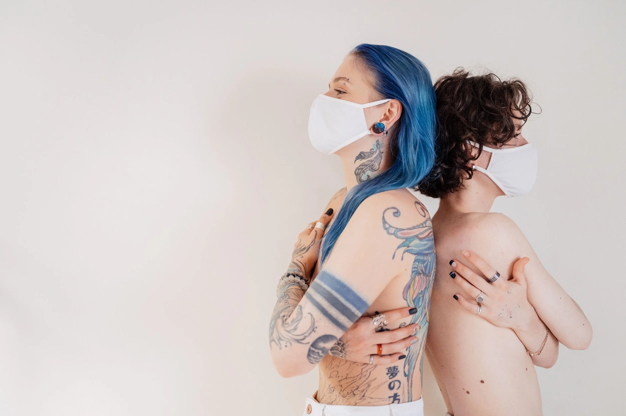 Two topless people back to back, hugging themselves, and wearing masks. One has long dark blue hair and tattoos, the other has chin length brown combination wavy curly hair. Both people have their nails done and are wearing multiple rings.