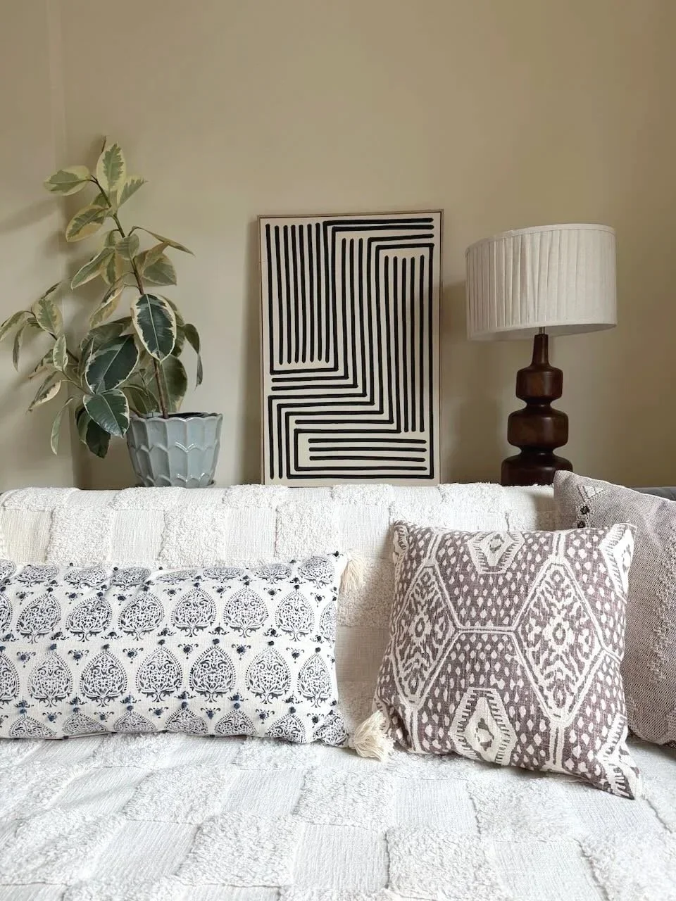 Living room corner with white sofa, patterned pillows, a potted plant, a black and white framed abstract art, a wooden table lamp with a white shade.