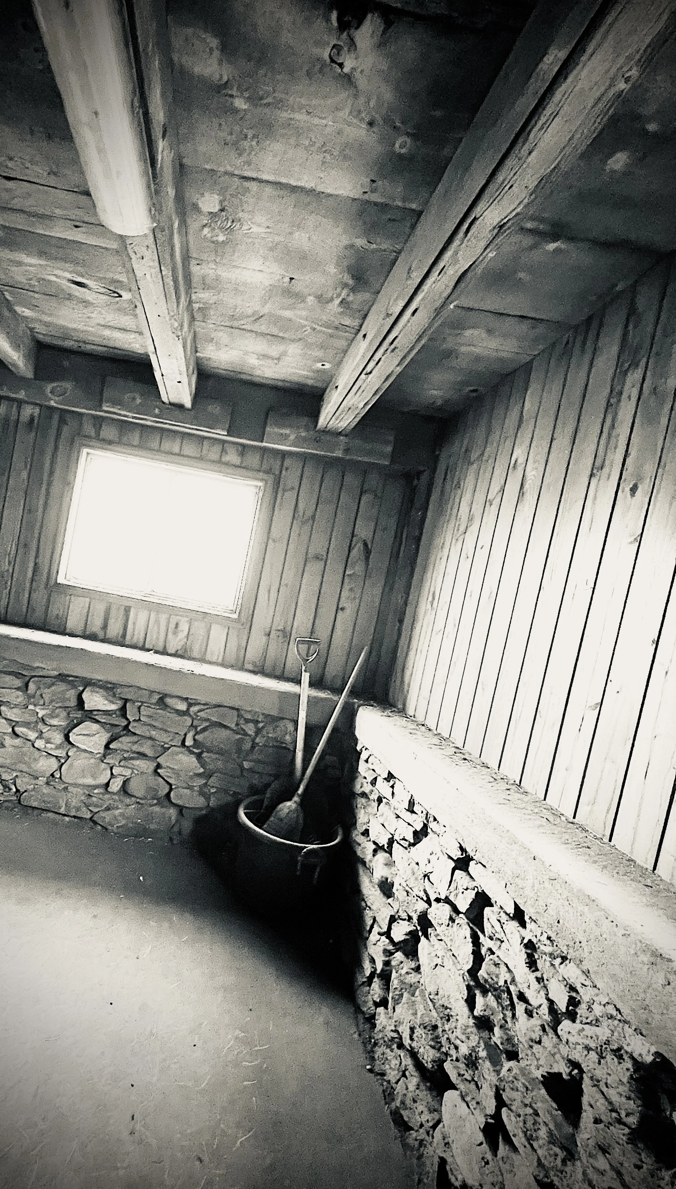Interior of a rustic room with wood-paneled walls and ceiling, a small window, a bucket, and a shovel leaning against the wall.