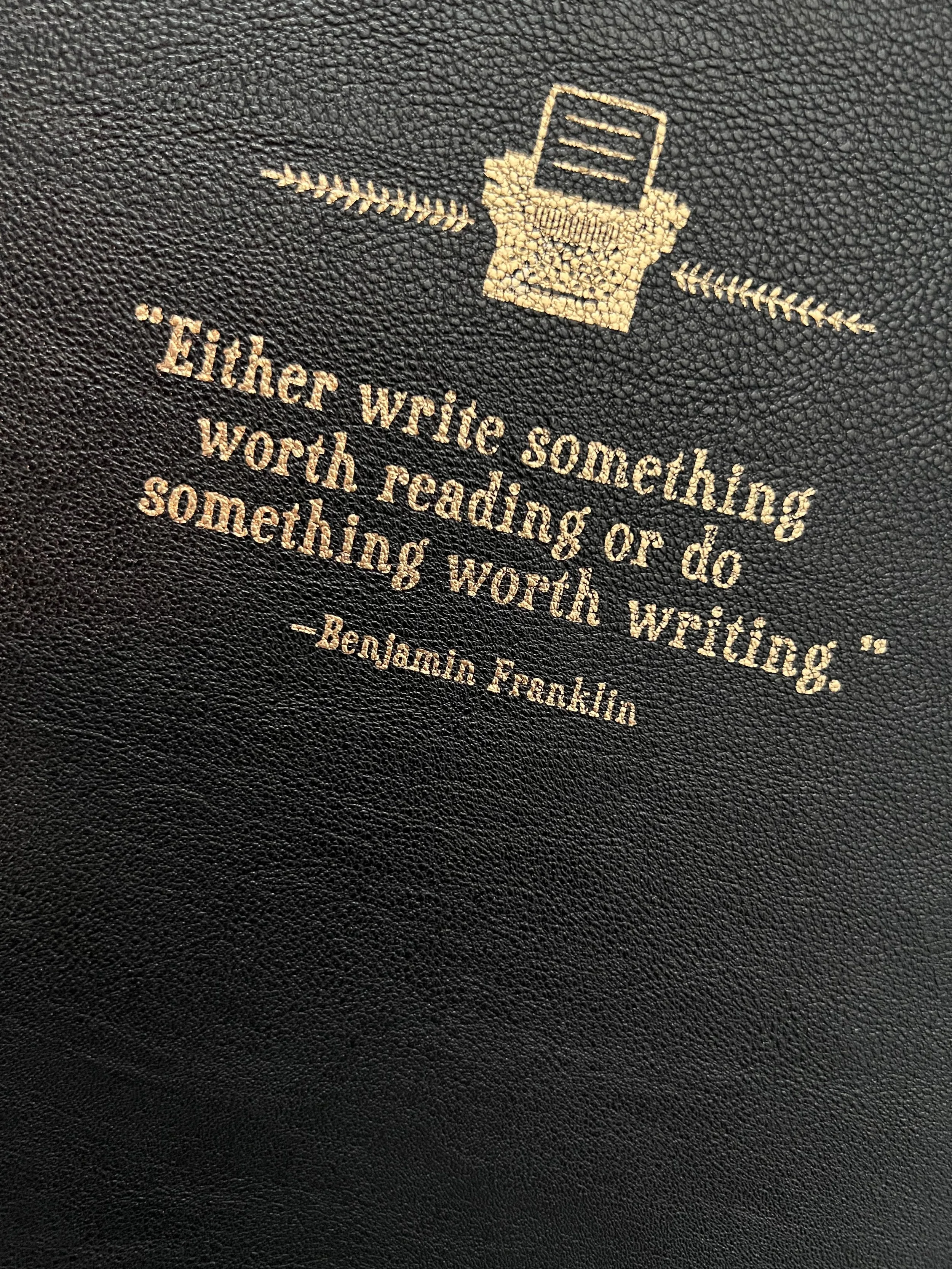 A black textured surface with a quote in gold lettering that reads: "Either write something worth reading or do something worth writing." -Benjamin Franklin. The quote is accompanied by a gold icon of a typewriter.